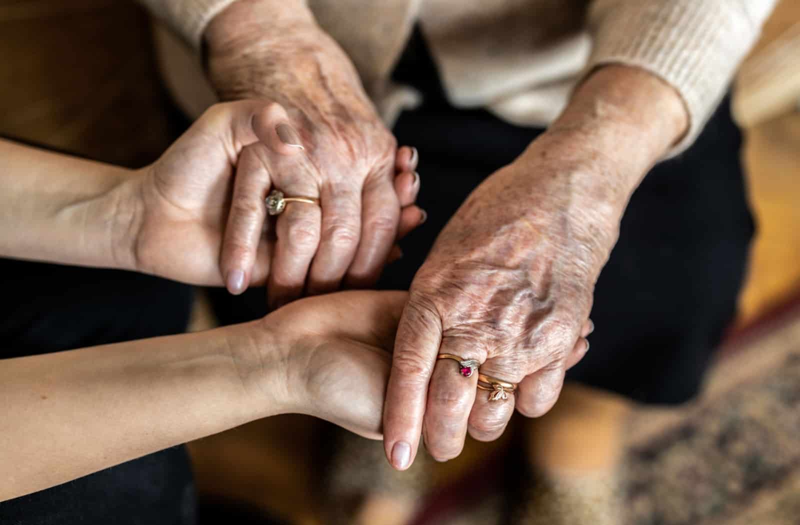 A middle-aged individual holds both of their senior parent’s hands to show their support