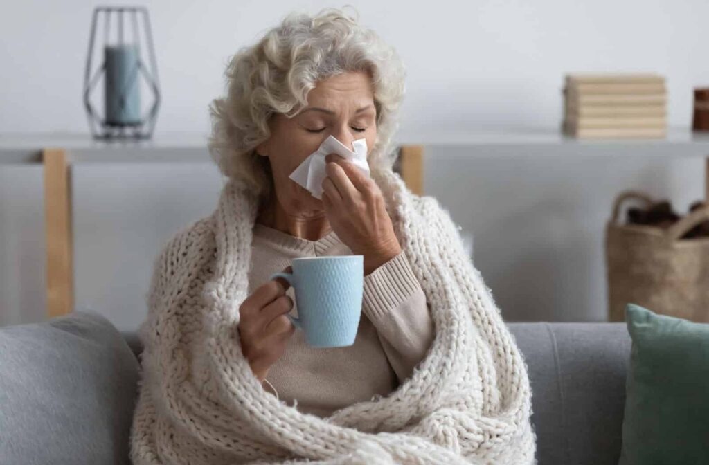 An older adult in a blanket blowing their nose and clasping a cup of tea to stop feeling so cold.
