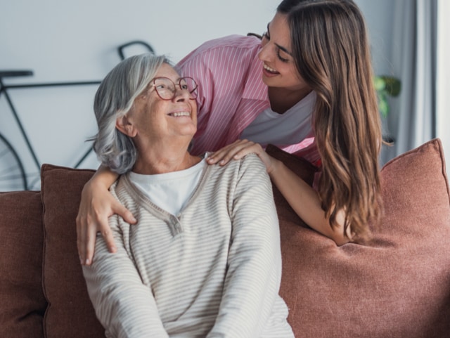 Young woman hugs an elderly woman on a sofa, both smiling warmly.