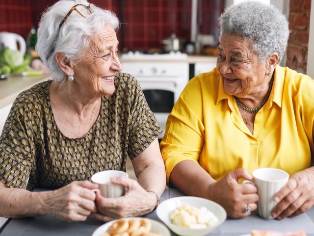 Two elderly women with gray hair sit at a kitchen table, smiling and chatting over mugs of tea or coffee with bowls of snacks nearby—a friendly, social moment in a home setting.