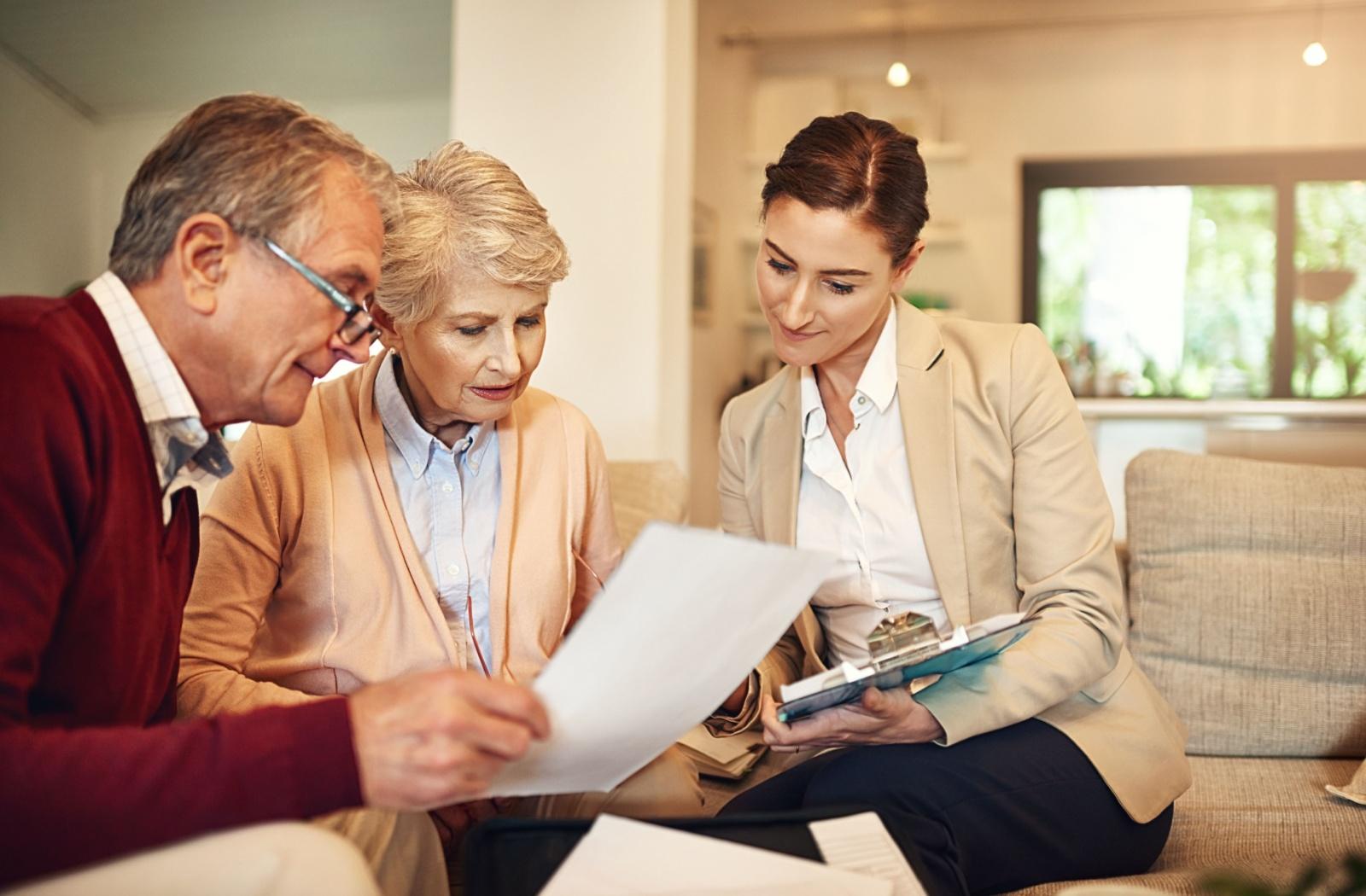 Three adults sit on a sofa, elderly couple reviewing papers with a younger professional. They look focused in a bright living room setting, with folders on their laps and a clipboard nearby.